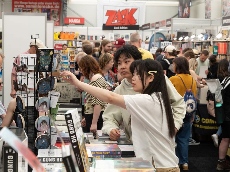 Eine belebte Comic-Convention mit zahlreichen Menschen an Ständen beim stöbern und Produkte wie Comics, Bücher und Merchandising-Artikel anschauen. 