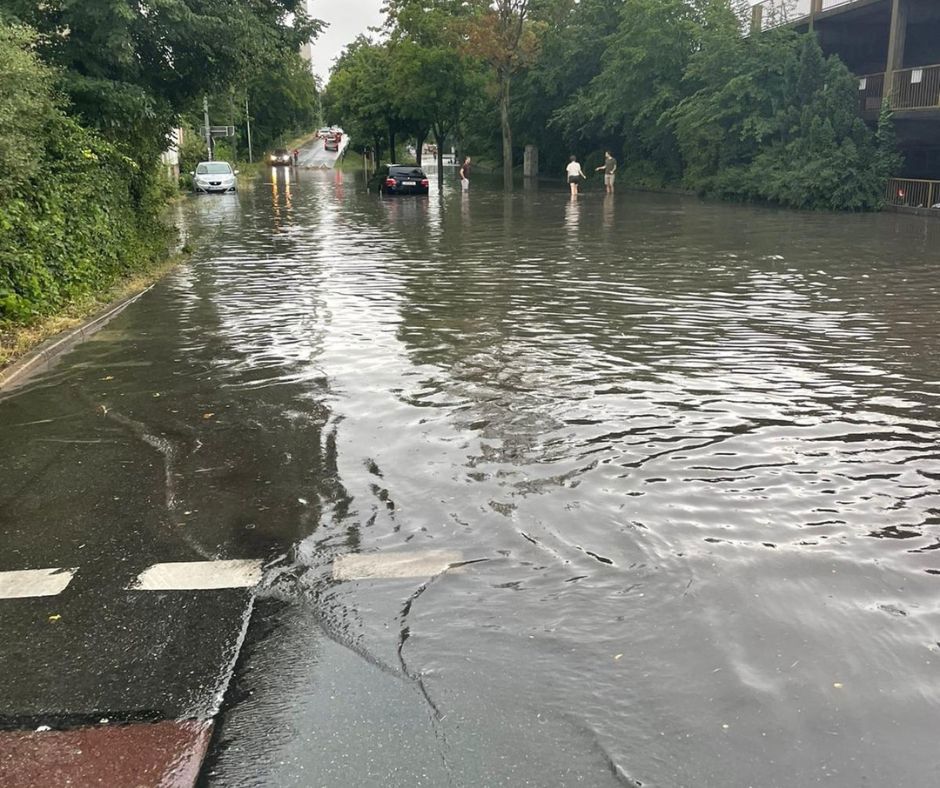 Eine Straße ist stark überflutet, mehrere Autos stehen im Wasser und einige Menschen waten durch das Hochwasser.