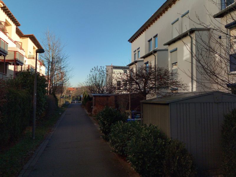 Ein schmaler Gehweg zwischen Wohnhäusern, gesäumt von Büschen und Bäumen mit einem klaren blauen Himmel im Hintergrund..