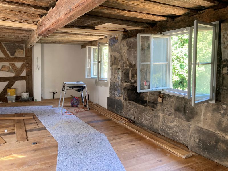 Innenraum mit Holzboden und Balken sowie zwei geöffnete Fenster.