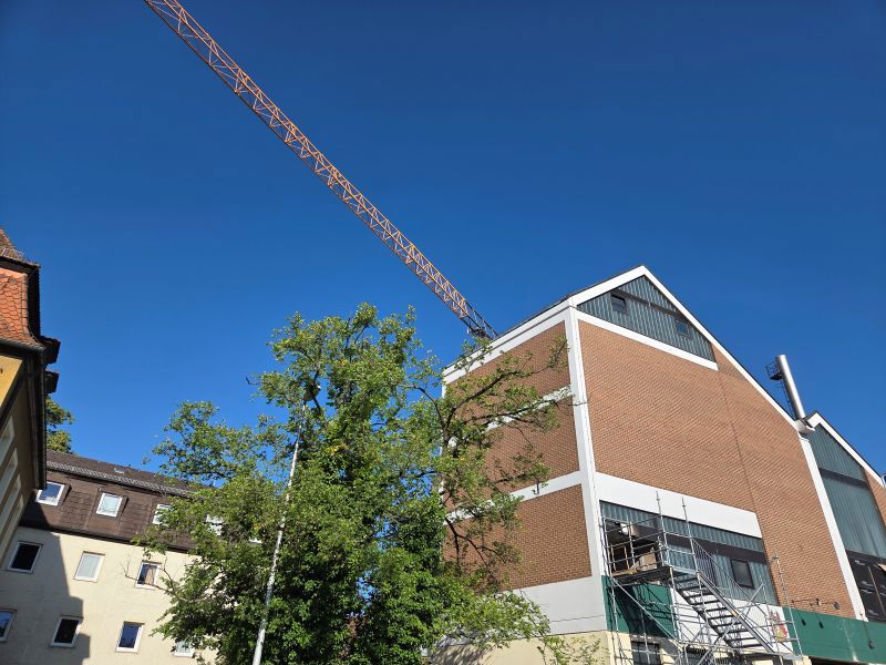 Vor blauem Himmel steht ein großes Industriegebäude mit rotbrauner Fassade und Baugerüst, daneben ragen grüne Bäume auf und ein Baukran erstreckt sich schräg über das Bild.