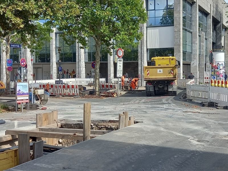 Eine Baustelle mit Absperrungen, einem gelben Baustellen-Lkw, mehreren Bauarbeitern in orangener Kleidung und einem offenen Straßenabschnitt. Im Hintergrund stehen moderne Bürogebäude und Bäume.