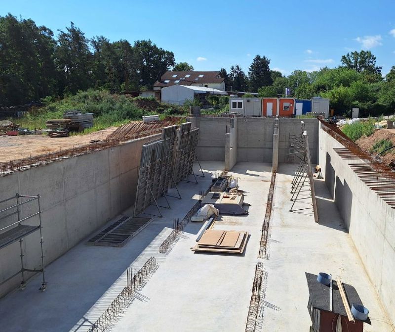 Eine große Baugrube mit bereits errichteten Betonwänden und verschiedenen Baumaterialien sowie Baugeräten im Inneren. Im Hintergrund steht ein Baucontainer und ein Wohnhaus vor einer grünen Baumkulisse.