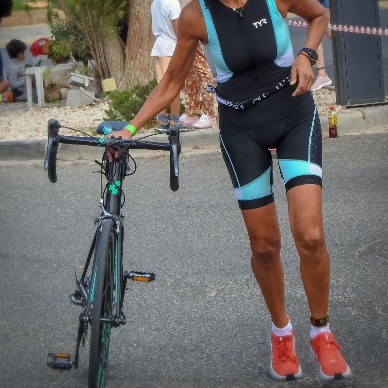 Eine sportlich gekleidete Frau in einem Triathlonanzug schiebt ihr Rennrad laufend über eine asphaltierte Straße, während im Hintergrund Zuschauer sitzen und der Bereich mit Absperrband markiert ist.