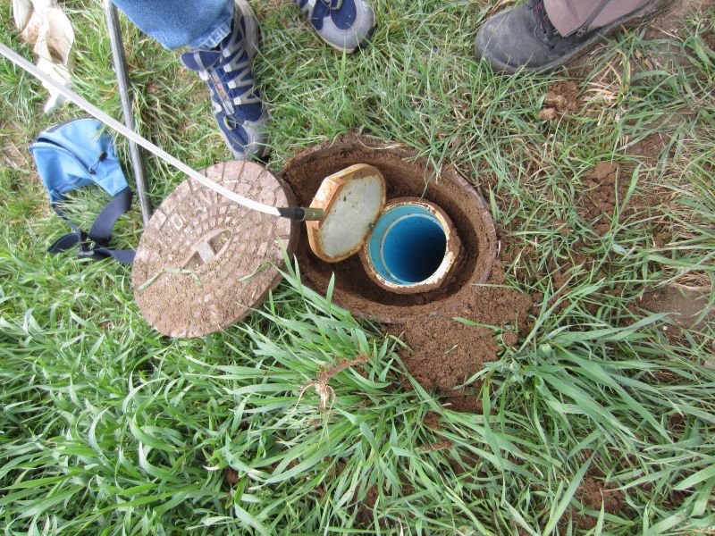 Auf dem Bild ist ein geöffneter Grundwassermessbrunnen auf einer Wiese zu sehen, aus dem mithilfe eines Schlauchs eine Wasserprobe entnommen wird; am Rand stehen Personen mit festem Schuhwerk.