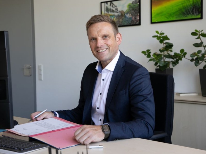 Oberbürgermeister Jörg Volleth im Anzug sitzt lächelnd an einem Schreibtisch, hält einen Stift in der Hand und arbeitet an einer Akte, während im Hintergrund Zimmerpflanzen und gerahmte Bilder an der Wand zu sehen sind.