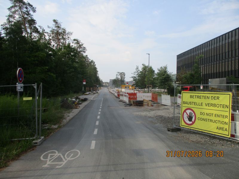 Eine gesperrte Straße mit Fahrradweg führt durch eine Baustelle, auf der Absperrzäune, Warnschilder und Baugeräte stehen. Zu sehen ist auch ein großes gelbes Schild mit dem Hinweis, dass das Betreten der Baustelle verboten ist.
