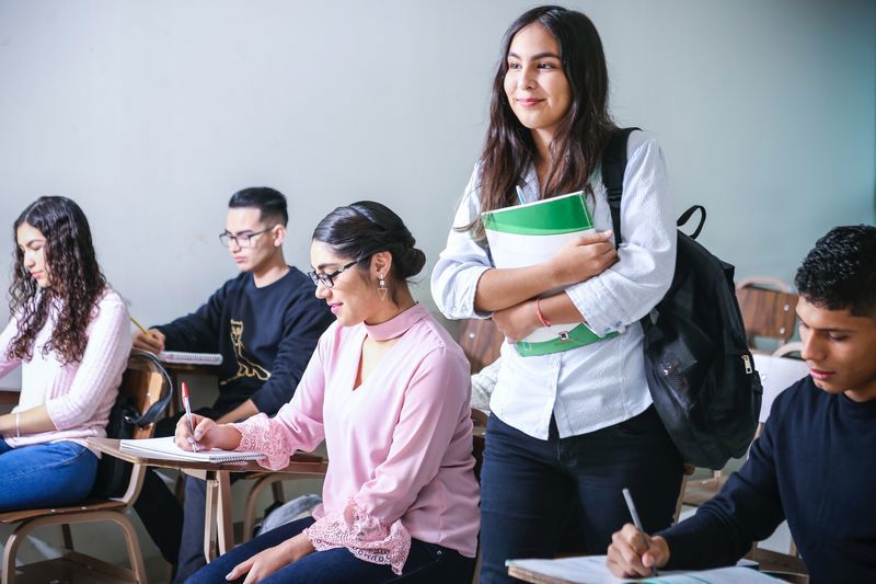 Klassenraum mit mehreren Schüler*innen. Eine Schülerin steht mit Rucksack und Buch unter dem Arm in der Mitte.