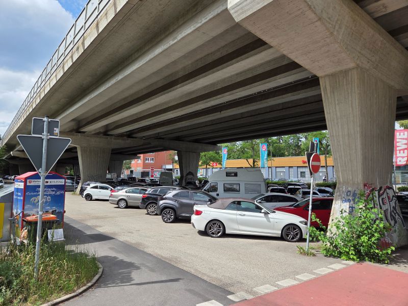 Unter einer großen Brücke befindet sich ein Parkplatz mit mehreren geparkten Autos, darunter ein weißes Cabrio im Vordergrund.