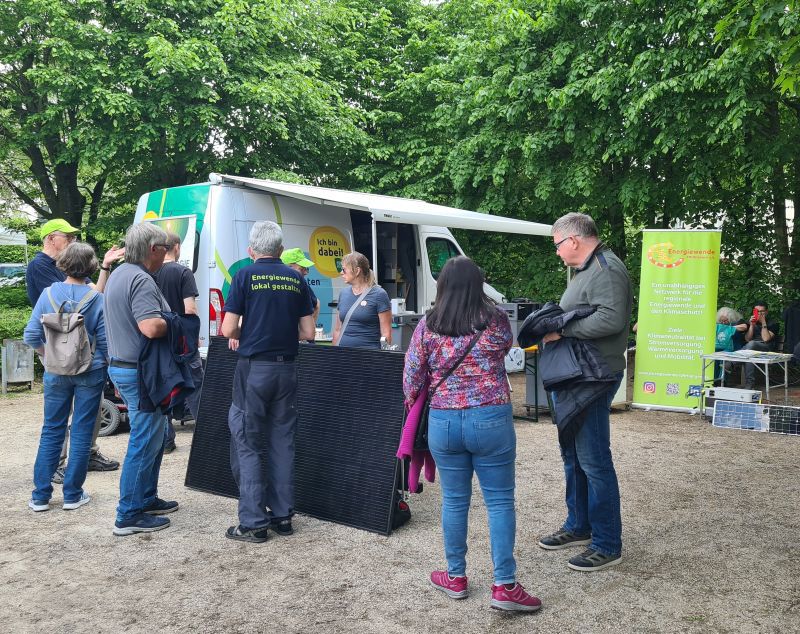 Mehrere Menschen stehen bei einem Informationsstand vor einem weißen Van im Freien, umgeben von Bäumen und Infotafeln.