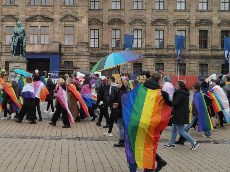 Eine Gruppe von Menschen läuft bei einer Demonstration durch eine Innenstadt und trägt dabei Regenbogenflaggen sowie andere Pride-Flaggen; einige halten einen bunten Regenschirm, im Hintergrund ist ein historisches Gebäude mit einer Statue zu sehen.