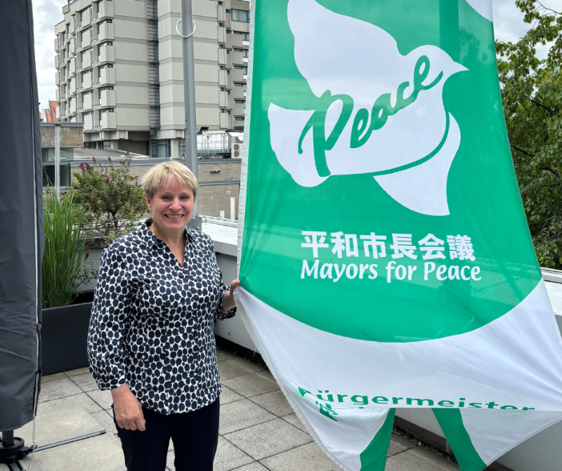 Eine lächelnde Frau steht auf einer Terrasse neben einer großen grünen Fahne mit einer weißen Friedenstaube und der Aufschrift 