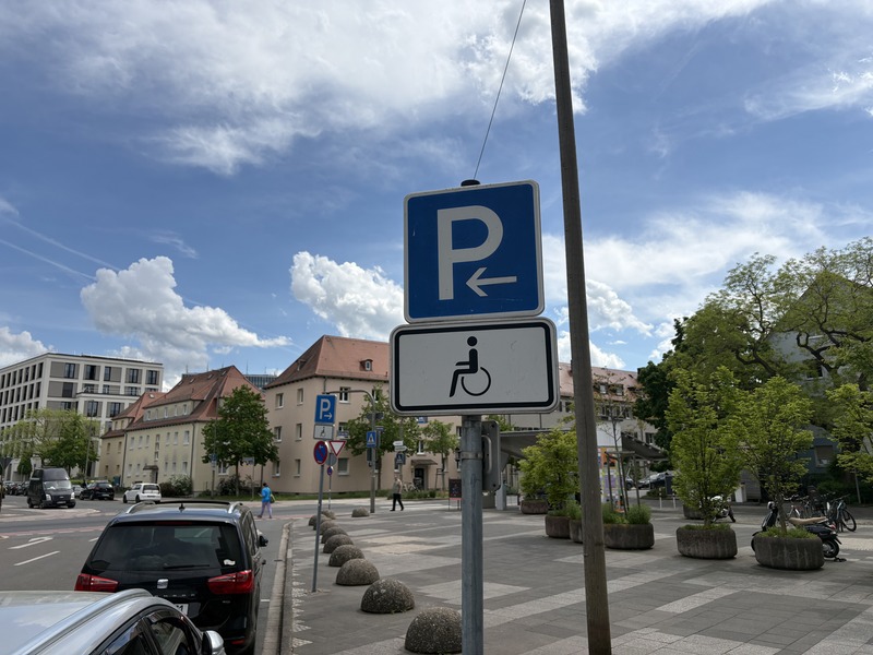 Ein blaues Parkplatzschild mit einem Pfeil nach links und einem darunter angebrachten Symbol für Behindertenparkplätze steht am Straßenrand in einer urbanen Umgebung bei blauem Himmel mit einigen Wolken.