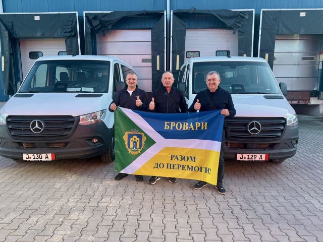Vasyl Dovgan, Leiter der Abteilung für Zivilschutz der Stadt Brovary  (links), Mykola Zinchenko, Stadtrat Brovarys (mitte) und  Petro Babych, stellvertretender Bürgermeister von Brovary (rechts) halten eine Fahne der Stadt Brovary hoch und zeigen einen Daumen hoch. Im Hintergrund stehen die zwei durch das Bundesministerium für Zusammenarbeit und der Gesellschaft für Internationale Zusammenarbeit geförderte Minibusse mit Rollstuhlrampe. 