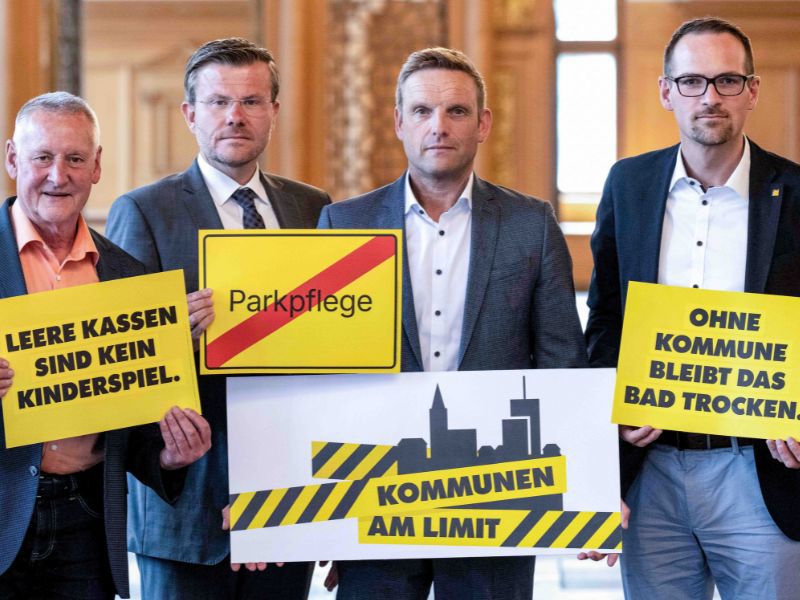 Vier Männer in Anzügen halten gelbe Protestschilder mit Aufschriften wie 