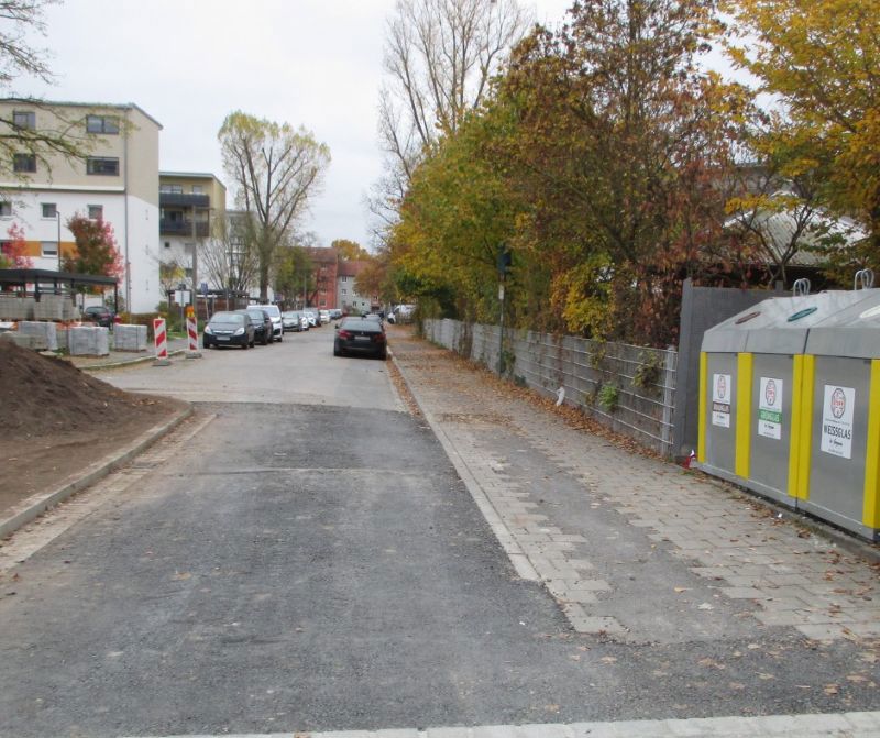 Eine teilweise neu asphaltierte Straße mit parkenden Autos, Baustellenmaterial, Altglascontainern und herbstlich gefärbten Bäumen am Straßenrand .