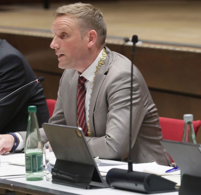 Oberbürgermeister Jörg Volleth im Anzug und mit Amtskette sitzt an einem Konferenztisch mit Mikrofon, Tablet und Getränkeflaschen.