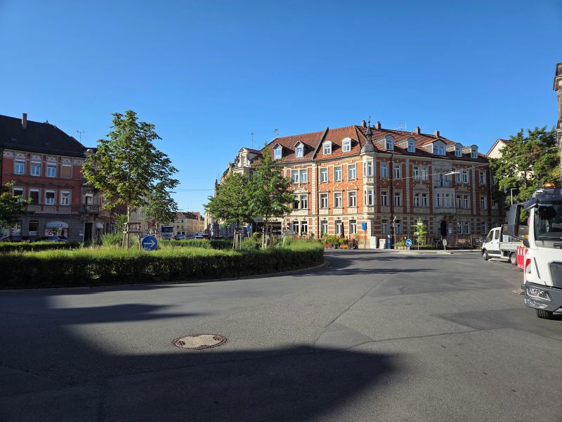 Ein kleiner Kreisverkehr mit begrüntem Mittelstreifen und jungen Bäumen vor einer historischen Häuserzeile mit roten Ziegeln und weißen Fensterrahmen. Am rechten Bildrand ein LKW und der Himmel strahlend blau.