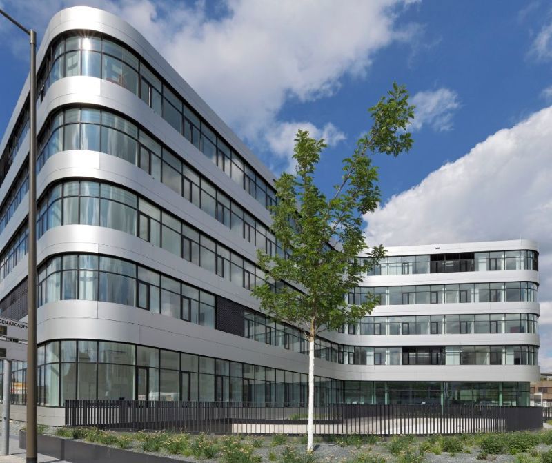 Ein modernes Bürogebäude mit abgerundeten Ecken und großen Glasfenstern steht unter blauem Himmel, während im Vordergrund ein junger Baum und eine kleine Grünfläche.