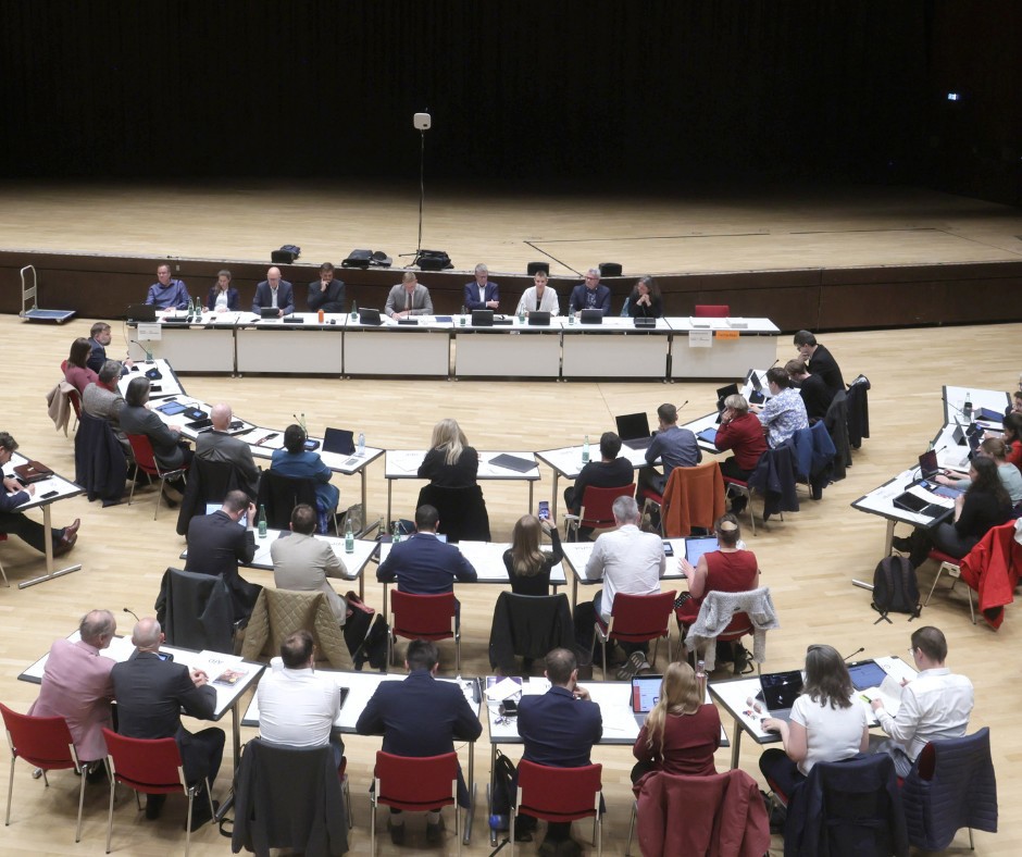 Eine größere Gruppe von Menschen, die in einem großen, halbrund angeordneten Sitzungssaal an Tischen sitzen.