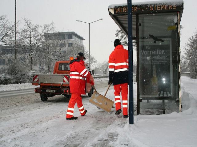 Zwei Mitarbeiter beim Schnee räumen vor der Bushaltestelle Werner-von-Siemens-Straße