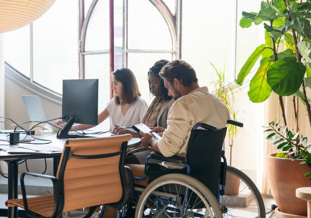 Drei Menschen, teilweise im Rollstuhl sitzend, arbeiten gemeinsam ein einem Schreibtisch
