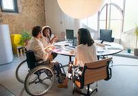 Man in a wheelchair sits at a table in an office with two women, they are talking