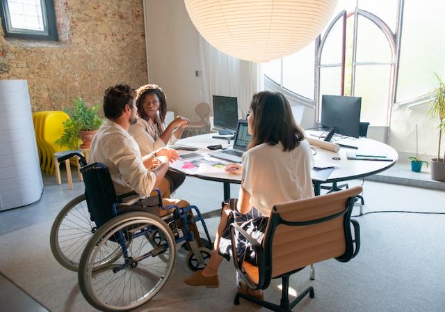 Mann im Rollstuhl sitzt mit zwei Frauen an einem Tisch im Büro, sie unterhalten sich
