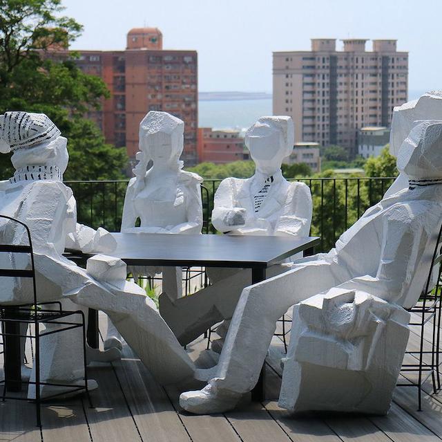 Five white stone figures sit around a table outside. They symbolically represent public cultural-political discourse.