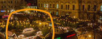 Ein stimmungsvoll beleuchteter Weihnachtsmarkt mit zahlreichen Ständen und vielen Besuchern erstreckt sich vor einem historischen Gebäude im Abendlicht; mittig oben ist ein Bereich digital überdeckt.